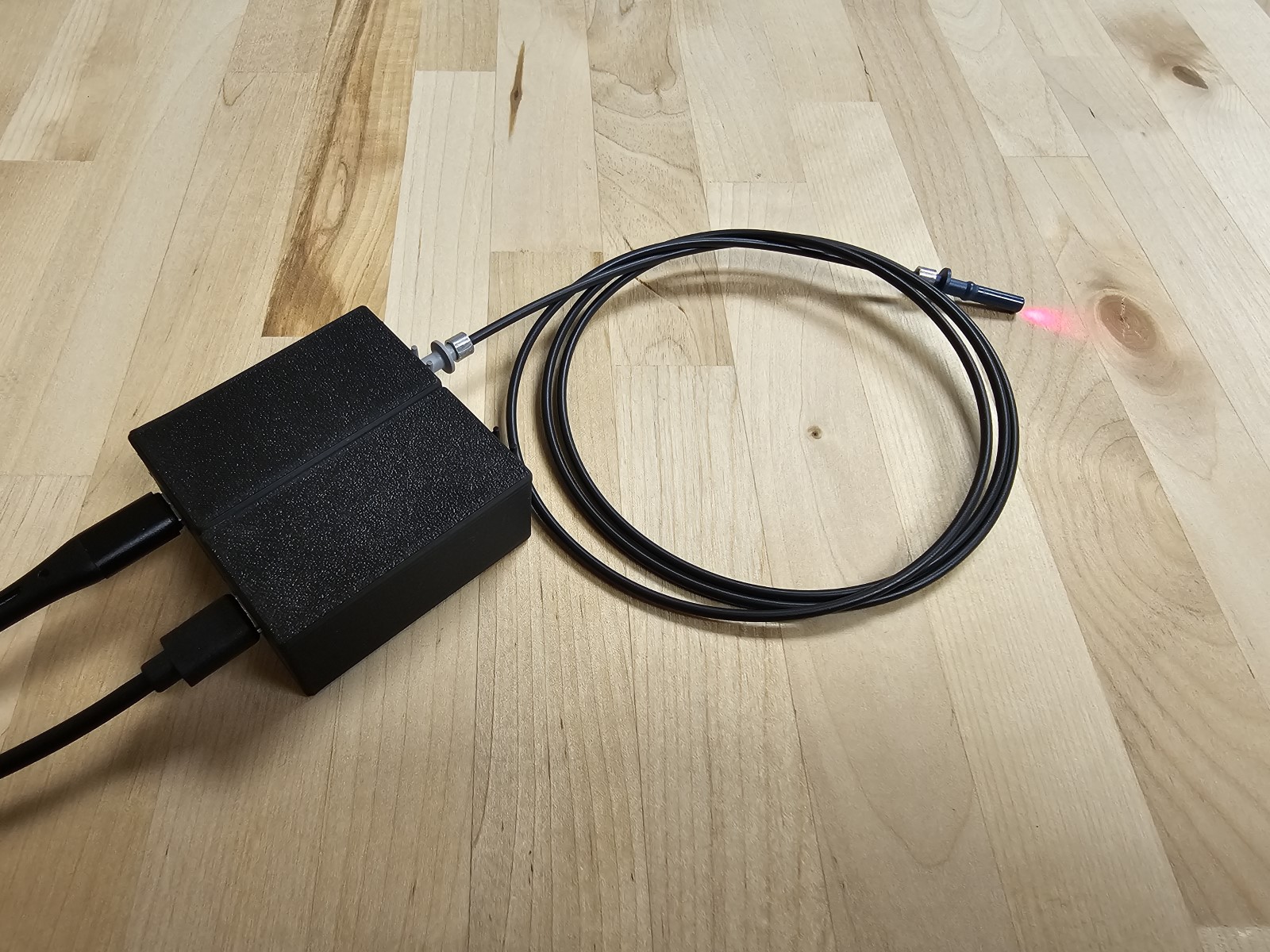 A transmitter enclosure with the fiber attached at its grey port. The
         far end of the fiber is unplugged, lying on the workbench, with
         visible red light coming out of it.