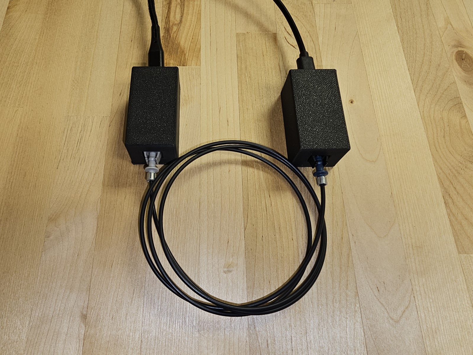 Two small black enclosures on a workbench, each with a USB cable
         running away to its own host. A single optical fiber, coiled between
         them, runs from the grey port on the left unit to the port on the
         right unit.