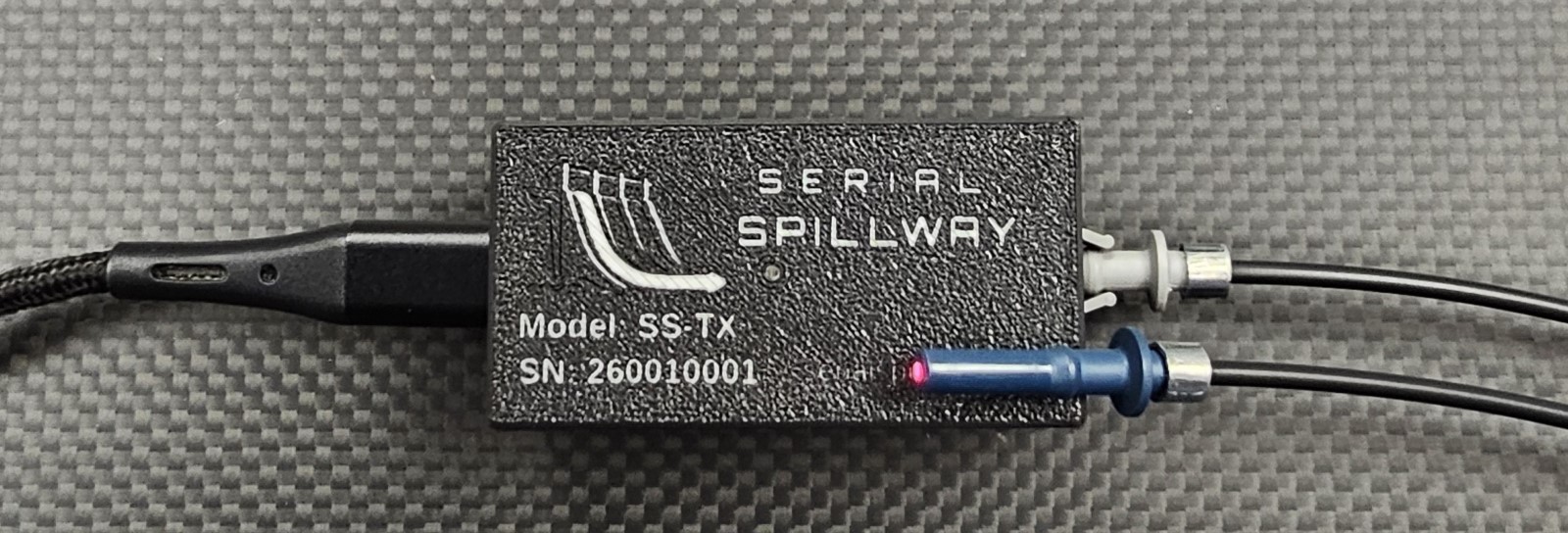 A transmitter unit, marked SERIAL SPILLWAY, Model SS-TX, with a
         serial number. A USB-C cable enters at one end. A single grey optical
         port carries the fiber. The fiber's other end, a blue connector,
         rests on top of the case and glows red.